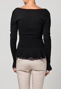 Nainen, jolla on yllään musta pitkähihainen istuva paita, jossa on röyhelöhelma, ja vaaleanbeige housut, näkyy selkäpuolelta yhtenäistä taustaa vasten.