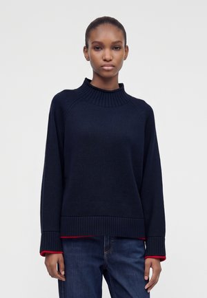 Femme portant un pull côtelé bleu marine foncé à col montant avec des poignets de chemise rouges visibles, associé à un jean bleu sur un fond uni.