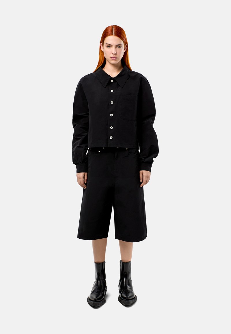 Zwarte cropped jack met grote kraag en knoopsluiting, gecombineerd met zwarte shorts tot de knie en zwarte enkelboots. Glad textuur, casual ontwerp.