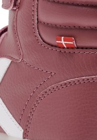 Profil latéral d'une chaussure en cuir rose avec des accents blancs, des coutures visibles et une petite étiquette rouge. Texture lisse avec un design structuré.