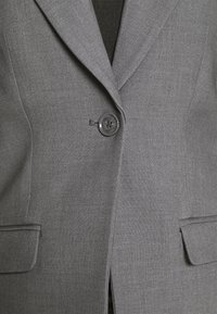 Blazer gris sur mesure avec un seul bouton foncé fermé, deux poches avant à rabat et un tissu noir visible sous les revers.
