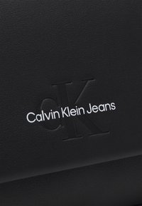 Materiale nero testurizzato con iniziali in rilievo "CK" e testo bianco "Calvin Klein Jeans" per il branding.