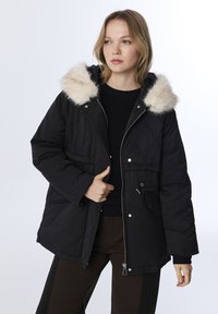 Zwarte gewatteerde jas met een met bont afgewerkt capuchon, ritssluiting, verstelbare taillekoord en voorkruiken, gedragen over een zwarte trui.