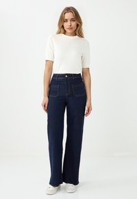 Jeans a vita alta blu scuro, larghi, con tasche frontali, cuciture a contrasto e chiusura con bottone, abbinati a un top attillato bianco a maniche corte.