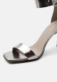 Ασημί μεταλλικό πέδιλο με ψηλό τακούνι, τετράγωνη μύτη, ένα μπροστινό λουράκι και αγκράφα στον αστράγαλο πάνω σε ανοιχτό φόντο.