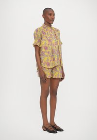 Gulvask med blomsterdesign og volanger på ermene, sammen med matchende shorts. Svarte flate sko har stropper og sirkulære detaljer.