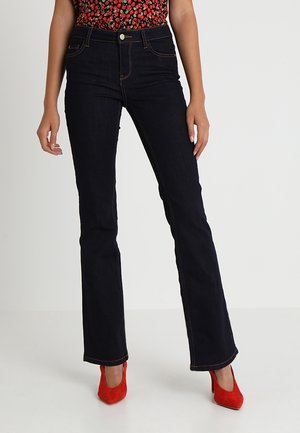 Kobieta ubrana w ciemnoniebieskie spodnie bootcut, czerwone buty na wysokim obcasie i czerwony wzorzysty top, stojąca na neutralnym tle.