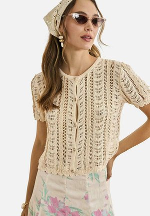 Mujer con top de manga corta de ganchillo color crema, falda floral, pañuelo beige de ganchillo en la cabeza, gafas de sol de ojo de gato y pendientes dorados de gota.