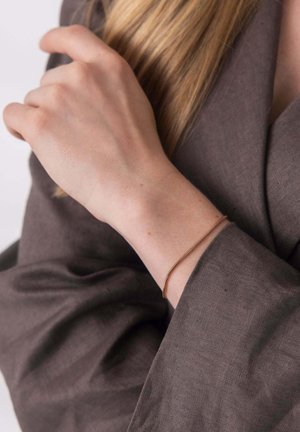 Dünnes goldenes Armband an einem Handgelenk, das gegen einen strukturierten Stoff in einem gedämpften Braunton ruht. Der Stoff hat ein weiches, natürliches Aussehen.