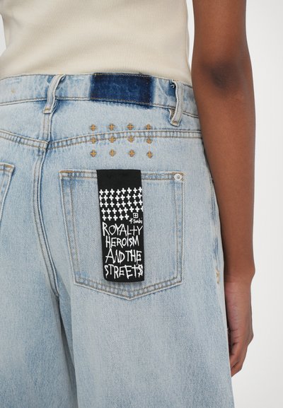 Lichtblauwe denim shorts met gouden kruissteek en een zwart label met witte tekst dat "ROYALTY HEROISM AND THE STREETS" bevat.