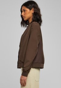 Giacca bomber marrone con una finitura liscia, polsini a coste e pannelli in rete sui lati per una maggiore texture. Chiusura con zip e design fitted.