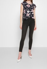 Femme portant un jean skinny noir, un haut à manches courtes fleurie bleu marine et des escarpins blancs à bride arrière se tenant sur un sol gris.