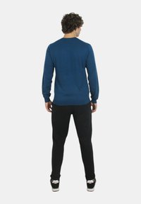 Maglione blu a collo rotondo realizzato in tessuto lavorato a maglia, abbinato a pantaloni neri. Modello visto da dietro, indossa scarpe casual con accenti bianchi.