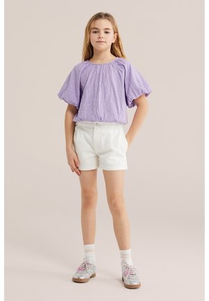 Fille debout, main dans la poche, portant un chemisier violet à manches courtes, un short blanc, des chaussettes blanches et des chaussures argentées avec des lacets roses.