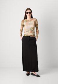 Long-sleeve fitted top with a postcard print in beige and brown, paired with a black maxi skirt. Model wears black sunglasses and flat shoes.