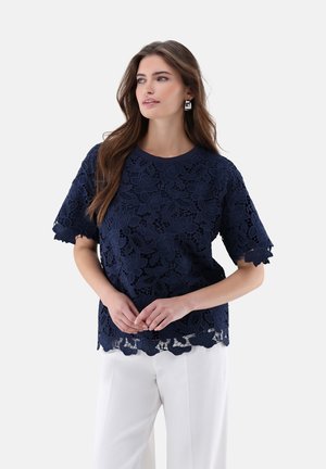 Donna che indossa una maglietta corta in pizzo floreale blu navy e pantaloni bianchi, posa con le mani leggermente incrociate, guardando leggermente verso destra.