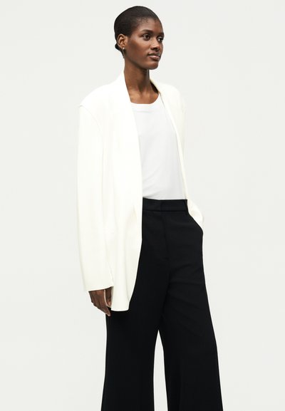 Γυναίκα με λευκό σακάκι και τοπ με μαύρο παντελόνι με φαρδιά μπατζάκια, στέκεται μπροστά σε απλό φόντο, με τα χέρια στις τσέπες, κοιτάζει μπροστά.