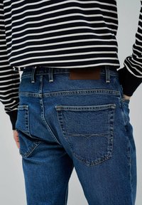Jeans de mezclilla oscuro con un corte clásico, con bolsillos traseros y una etiqueta de cuero marrón. Una camisa a rayas blanca y negra es parcialmente visible.
