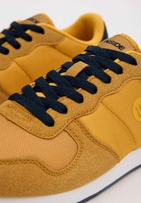 Zapatillas amarillas que combinan gamuza texturizada y malla suave, cordones azul marino y una suela blanca con un acento azul. Logo en el lateral.