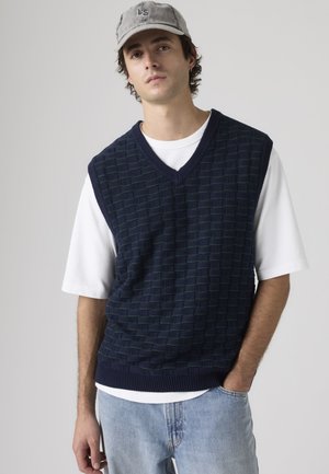 Tânăr cu vestă neagră tricotată texturată peste tricou alb, blugi albaștri deschis și șapcă gri, stând cu o mână în buzunar.