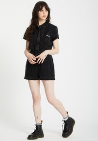 Mono corto de mezclilla negra con mangas cortas y cintura ajustada, que presenta rayas verticales y un pequeño logo. Combinado con botas negras hasta el tobillo y calcetines a rayas.