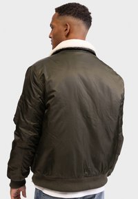 Olivově zelená bomber bunda s plyšovým bílým límcem, elastickými manžetami a spodním lemem. Disponuje hladkým, matným povrchem a kapsou na rukávu.
