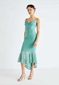 Rochie ajustată de culoare teal, cu un bodice din dantelă, decolteu adânc și un tiv evazat din dantelă. Prezintă bretele subțiri și un croi asimetric.
