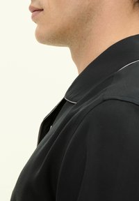 Maglietta nera con colletto, realizzata in un tessuto liscio, caratterizzata da una sottile profilatura grigia lungo il colletto. Vista di lato, che mette in mostra il collo.