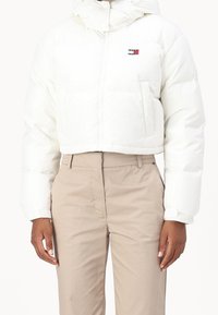 Vit kort vadderad jacka med huva, med en logotypmärke på bröstet och elastiska ärmslut. Bärs med beige byxor.