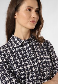 Ein Hemd mit Knopfleiste, das ein navy- und weißes abstraktes Muster aufweist, mit einem spitzen Kragen und schwarzen Knöpfen. Glatte Stofftextur.