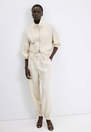 Mujer de pie contra una pared blanca, vestida con una chaqueta crema con cremallera y mangas remangadas, pantalones plisados a juego y zapatos marrones con punta abierta.