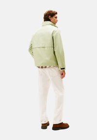Chaqueta verde claro con cuello alto, diseño ajustado y pantalones blancos. Zapatos marrones con suelas negras. Camisa a cuadros asoma por el dobladillo.