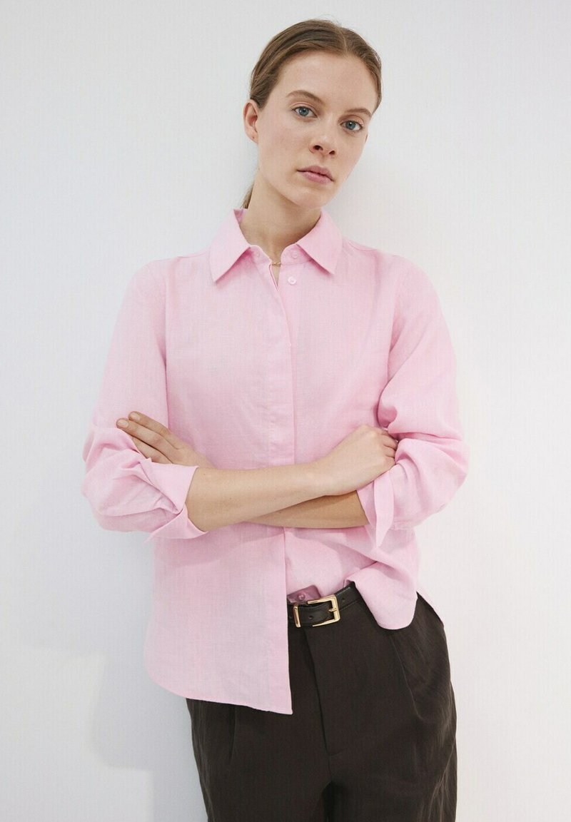 Frau mit hellbraunen Haaren steht mit verschränkten Armen, trägt ein locker sitzendes rosa Hemd zum Knöpfen und dunkle Hosen mit schwarzem Gürtel.