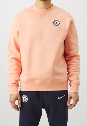 Homme portant un sweat-shirt pêche et un pantalon bleu marine avec les logos du club de football Chelsea et de Nike visibles sur les deux vêtements.