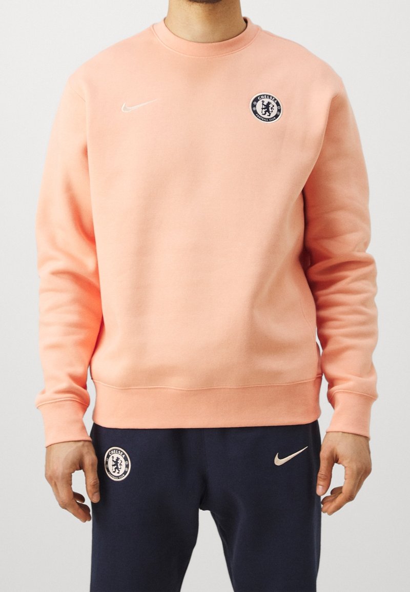 Homme portant un sweat-shirt pêche et un pantalon bleu marine avec les logos du club de football Chelsea et de Nike visibles sur les deux vêtements.
