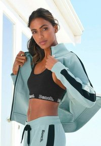 Hellblaues Trainingsanzug mit schwarzen Akzenten; Zip-Jacke kombiniert mit einem schwarzen Sport-BH und passenden Jogginghosen; Logo "Bench." auf dem Bund.