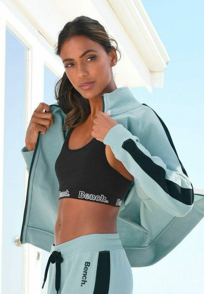 Hellblaues Trainingsanzug mit schwarzen Akzenten; Zip-Jacke kombiniert mit einem schwarzen Sport-BH und passenden Jogginghosen; Logo "Bench." auf dem Bund.