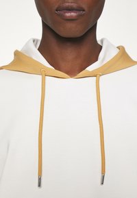 Huvtröja med en vit kropp, gula detaljer på huven och gula snören med silverfärgad spets. Slät textur.