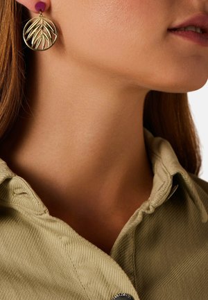 Profil d'une femme portant une boucle d'oreille circulaire en or avec un motif de feuille et une chemise beige à col.