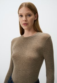 Top marrone a maniche lunghe realizzato in un tessuto morbido e testurizzato con scollatura a girocollo. Colore della pelle visibile attraverso il materiale, design aderente.