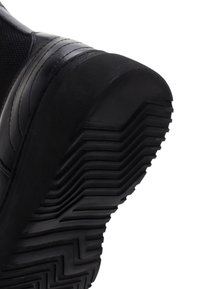 Semelle de chaussure de sport noire avec une surface en caoutchouc texturé. Présente des crampons épais et marqués, ainsi que des accents courbes lisses le long du bord.