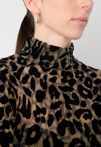 Leopardprint coltrui van doorschijnend materiaal in zwart en olijfgroen; met een hoge kraag en een textuurpatroon. Zilveren oorbellen in geometrische vorm.