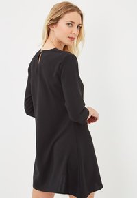 Mujer con cabello claro vistiendo un vestido negro de manga larga, por encima de la rodilla, girada parcialmente, mirando por encima del hombro contra un fondo blanco.