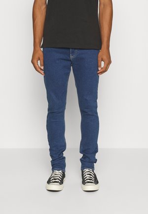 Jeans de mezclilla azul ajustados con una textura suave, que cuentan con un diseño clásico de cinco bolsillos y detalles de costura, combinados con zapatillas negras.
