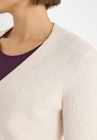 Cardigan beige claro con una superficie texturizada y un diseño de escote en V profundo. Ribete acanalado a lo largo del escote y una suave capa interior morada visible.
