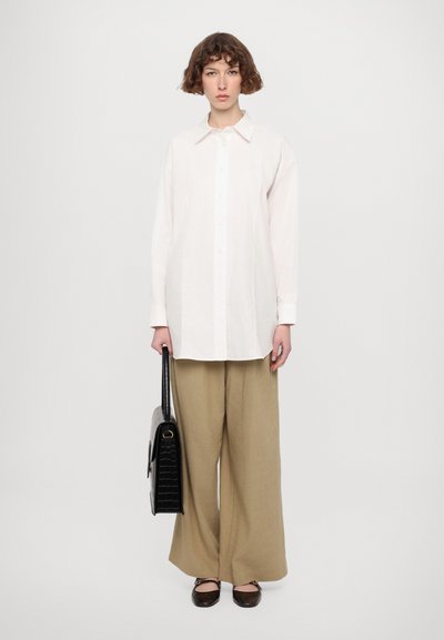 Camicia bianca a maniche lunghe, pantaloni beige larghi e scarpe basse marroni. Porta una borsa nera con texture e forma strutturata.
