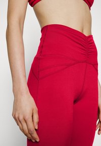 Vysoké červené legíny s nařaseným designem, vyrobené z elastického materiálu, s bočními švy a hladkou texturou.