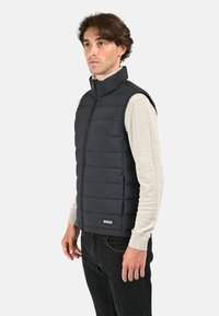 Gilet rembourré noir avec un col haut, fermeture éclair frontale et accent de logo. Porté par-dessus une chemise à manches longues gris clair et associé à un jean foncé.