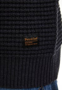 Gebreide trui in donker marineblauw met een textuurpatroon. Heeft een rib geborduurd rand en een zwart label met oranje tekst dat "Petrol Ind." vermeldt.