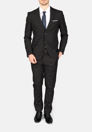 Man staat met een zwart pak, wit overhemd, donkerblauwe stropdas, zwarte nette schoenen en een wit pochet op een lichte achtergrond.